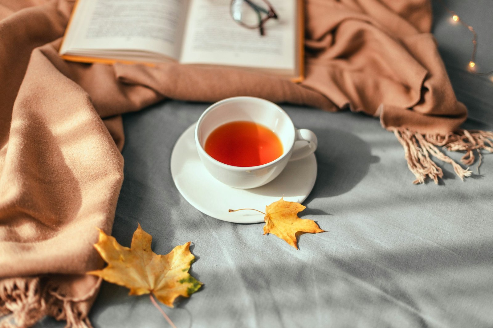 Warm knitted beige scarf, a cup of hot tea and an open book in bed. Cozy autumn